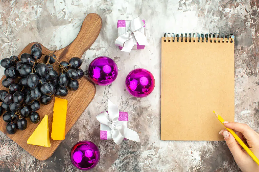 vista-superior-del-delicioso-racimo-de-uva-negra-fresca-y-queso-en-la-tabla-de-cortar-de-madera-y-accesorios-de-decoracion-de-regalos-y-escritura-mano-en-el-cuaderno-sobre-fondo-de-color-mezclado